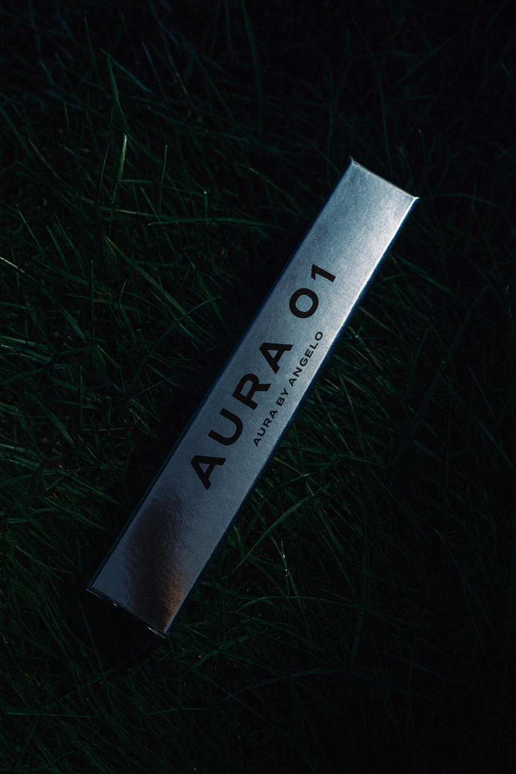 Aura 01 Fragrance silver packaging on dark grassy backdrop. 