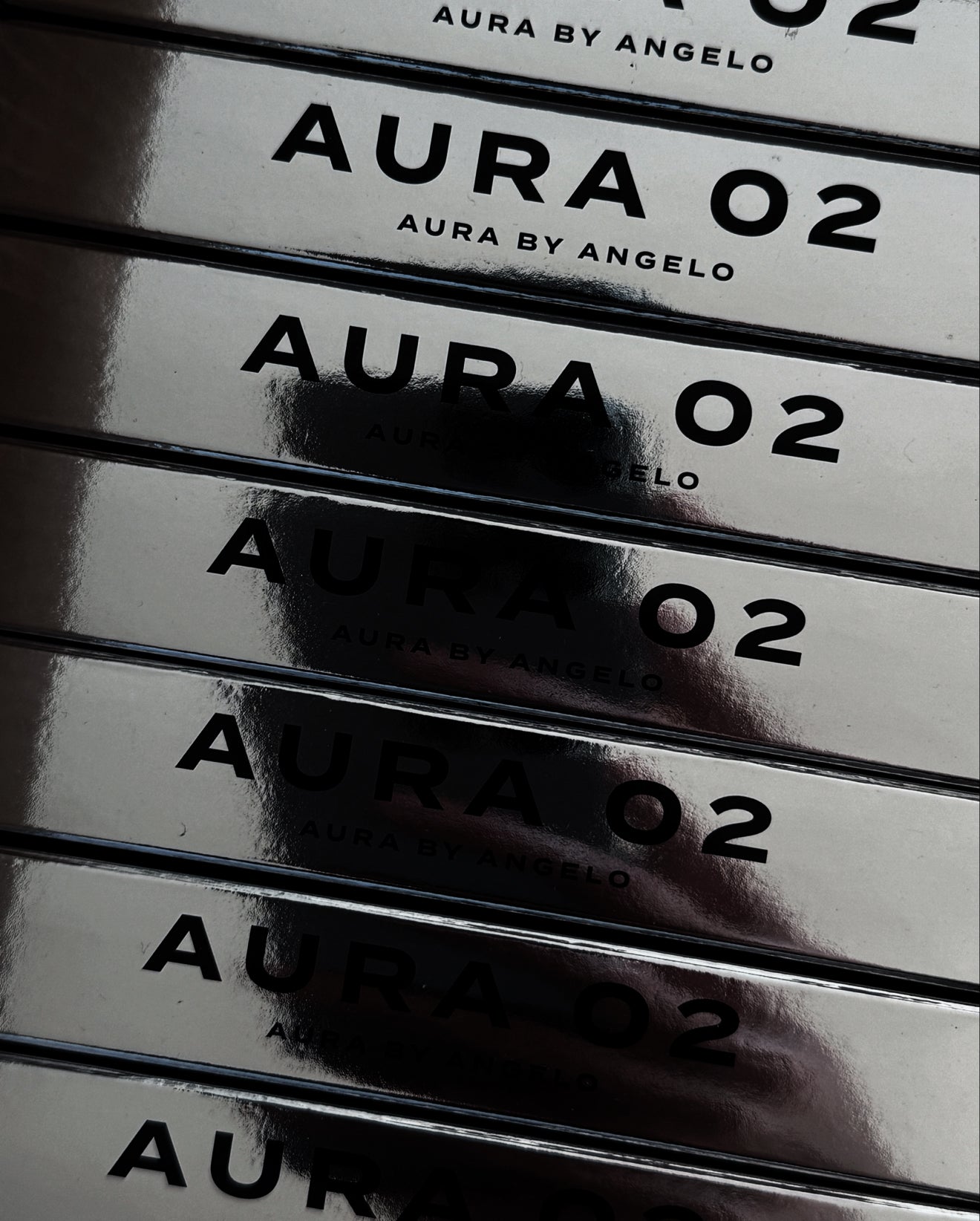 Stack of AURA 02 fragrance boxes showcasing a reflection of a hand on the silver packaging.  