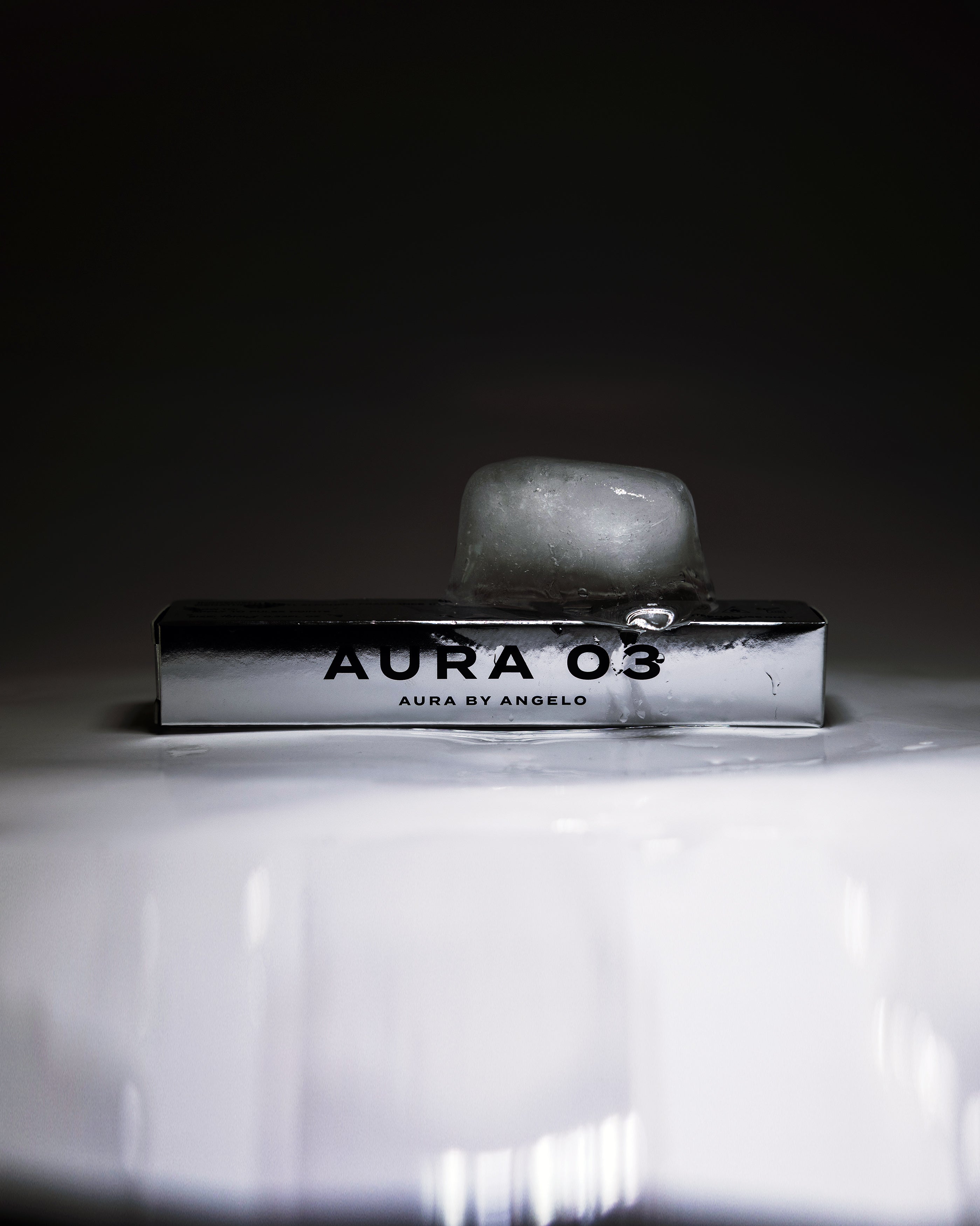 An ice cube melting on a metallic product box with 'Aura 03' fragrance branding against a dark background. 