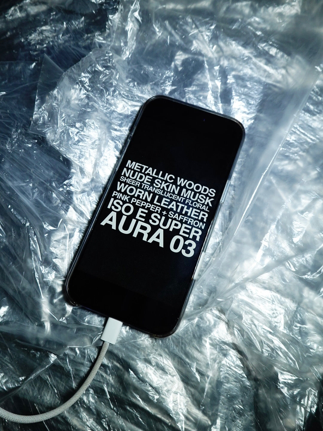 Phone with text on screen showcasing fragrance notes "Metallic woods, nude skin musk, sheer translucent florals, worn leather, pink pepper + saffron, iso e super, aura 03" against a textured background. 