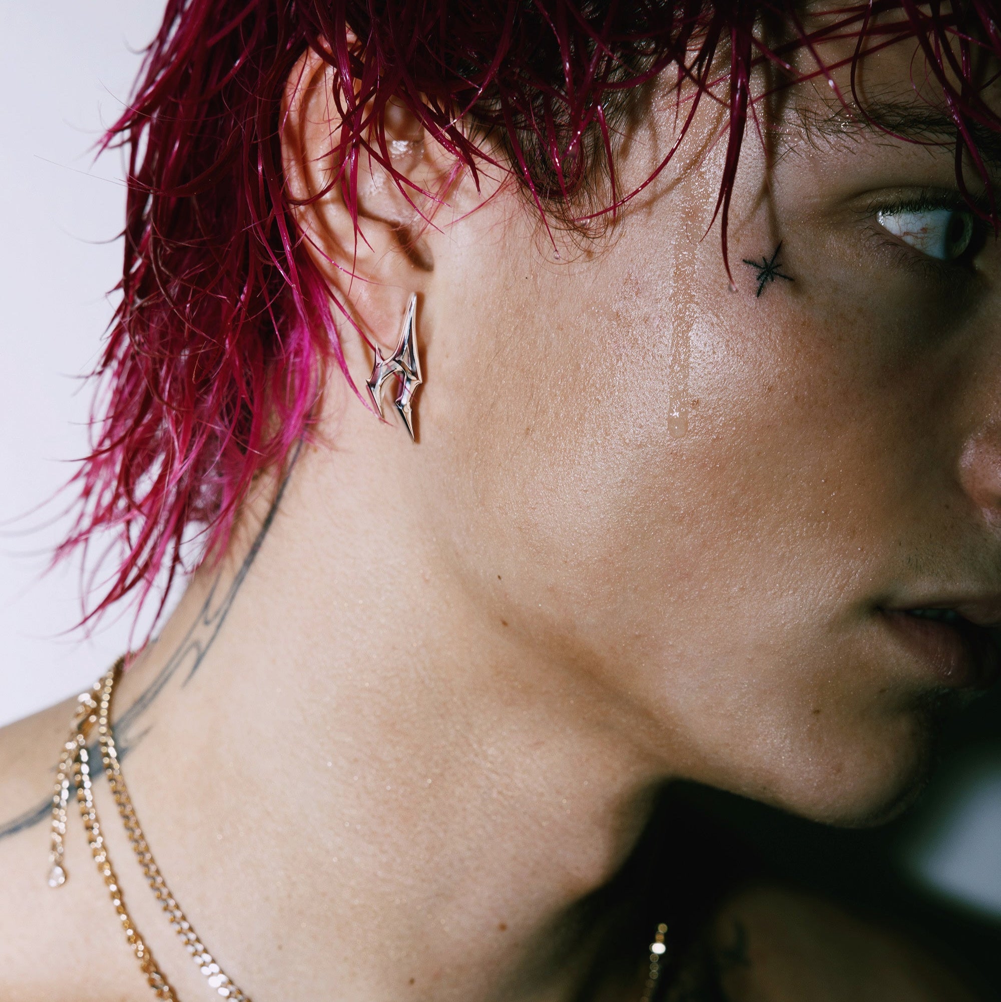 Male model with pink hair wears a silver metallic earring in the shape of an A from the AURA BY ANGELO logo. Drip of sweat comes down his face and accentuates his face tattoos. 
