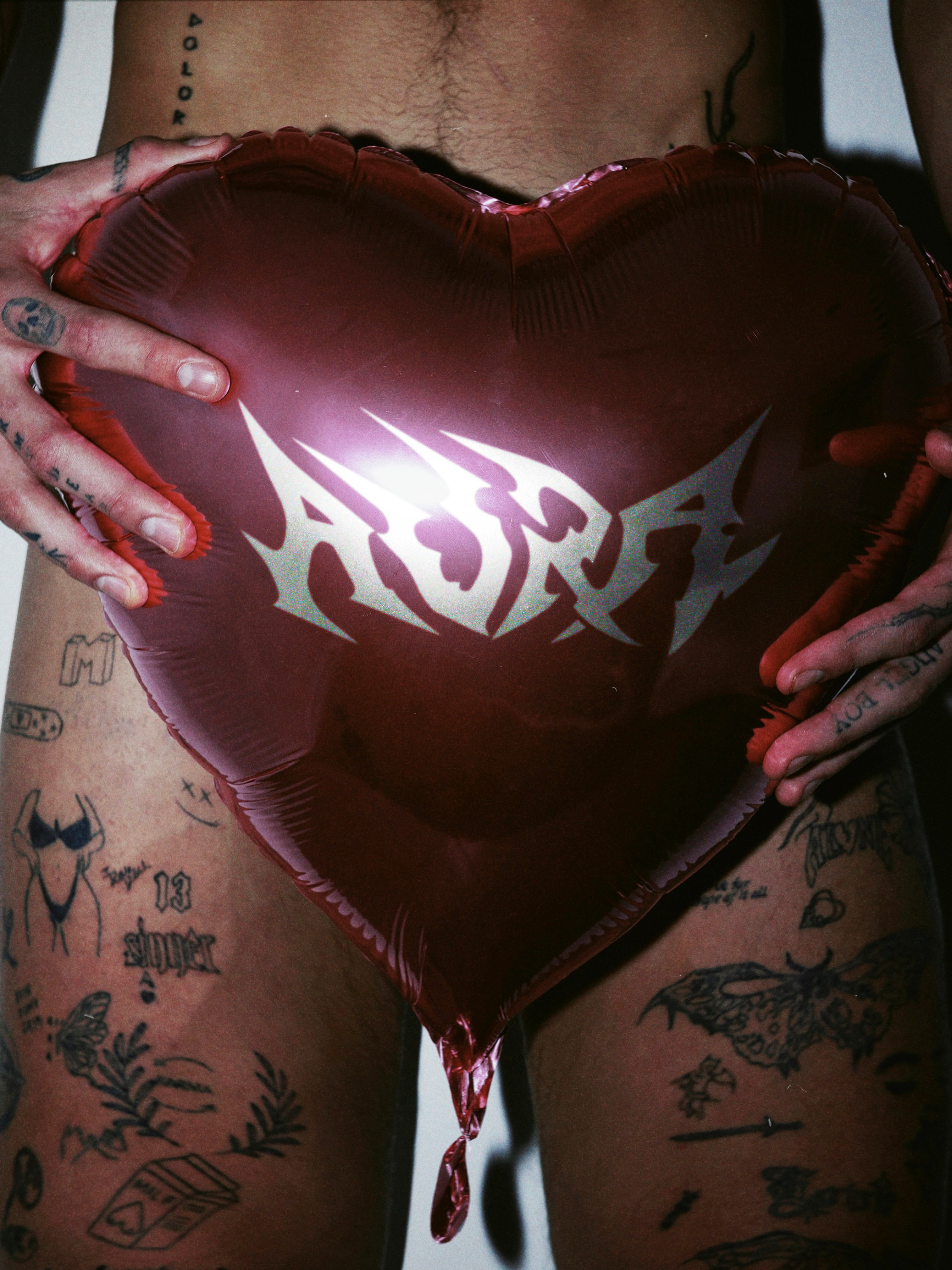 Close up of a man with tattoos holding a red heart shaped balloon with the AURA BY ANGELO logo on it in silver. 