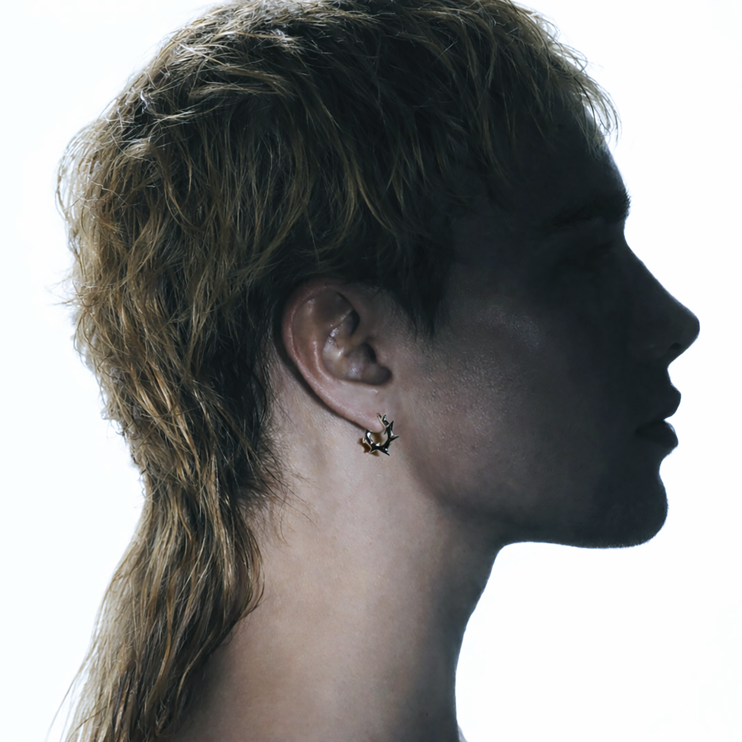 Silhouette profile of a blonde male model wearing a silver spiked hoop earring with a white background. 