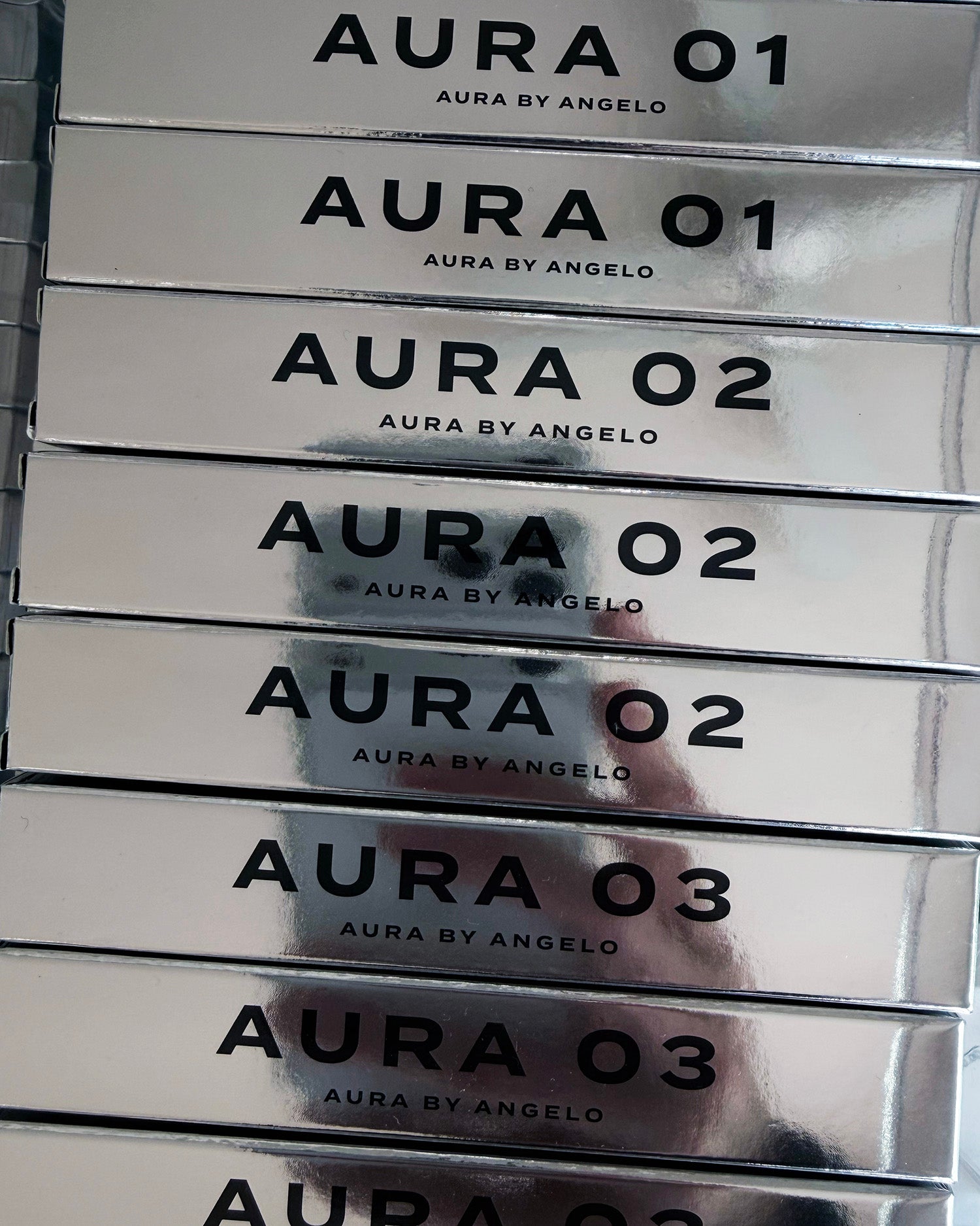 Stack of AURA BY ANGELO fragrances with reflection of hand photographing the mirror-like product boxes. 