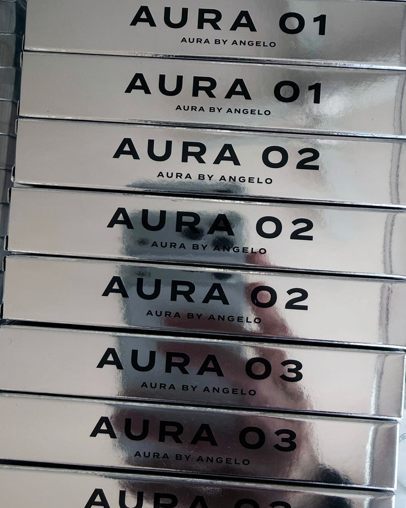 Stack of AURA BY ANGELO fragrances with reflection of hand photographing the mirror-like product boxes. 