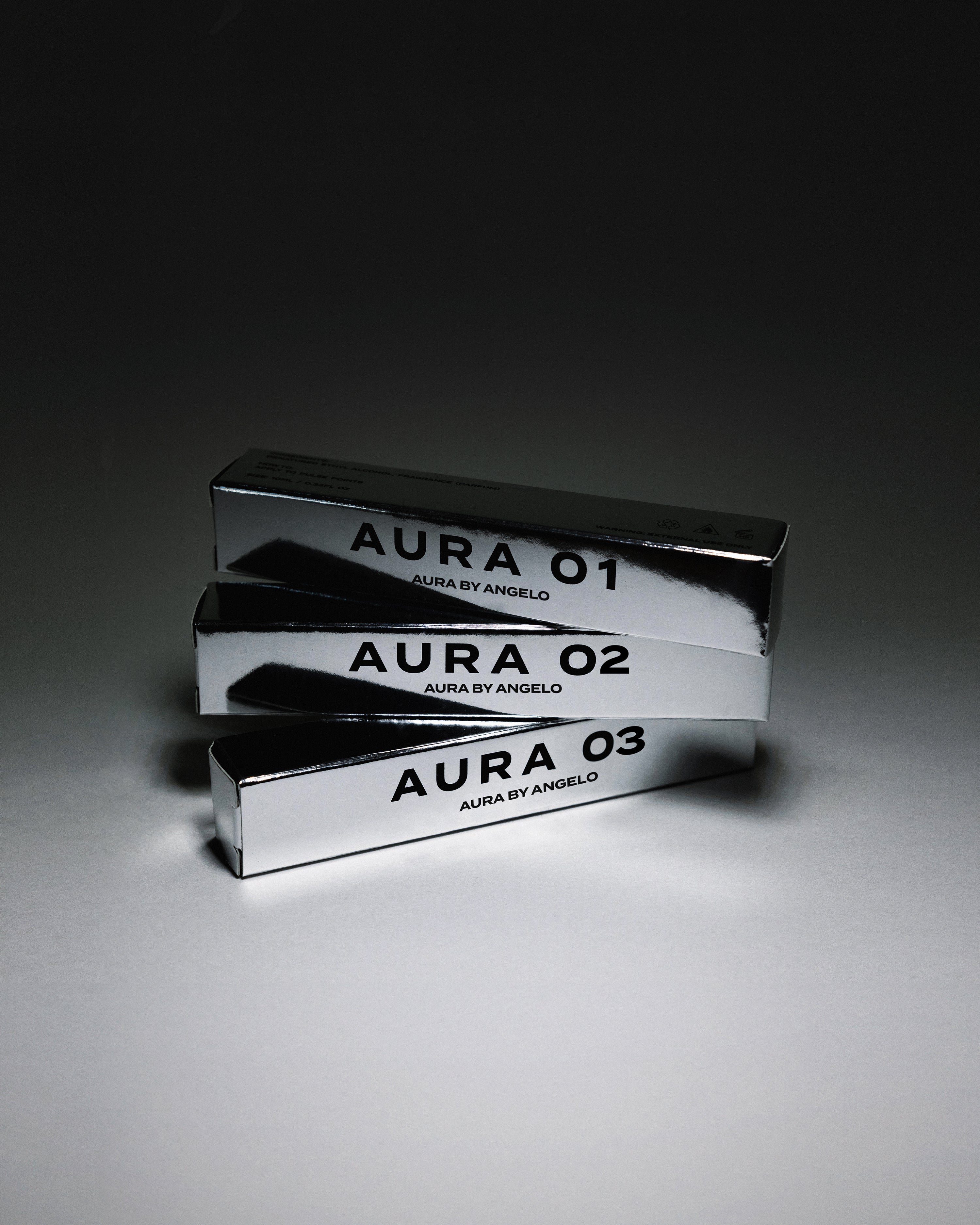 Three metallic fragrance boxes labeled 'Aura 01', 'Aura 02', and 'Aura 03' on a gray background.